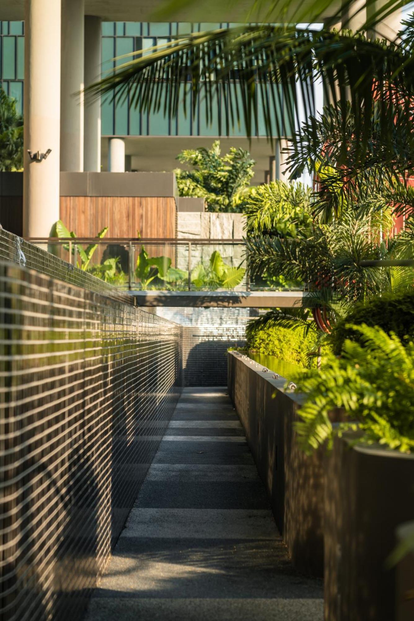 Parkroyal Collection Pickering, Singapura Luaran gambar The photo depicts a modern outdoor space featuring sleek design elements. In the foreground, there is a narrow walkway bordered by reflective surfaces. On one side, lush green plants and ferns are arranged in a planter, adding a natural touch to the 