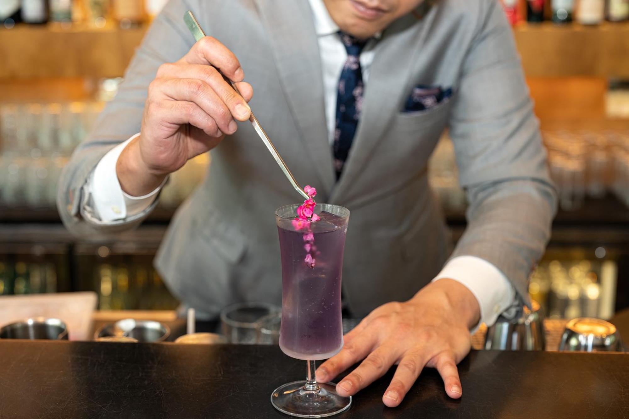 Parkroyal Collection Pickering, Singapura Luaran gambar The photo shows a bartender in a stylish gray suit preparing a cocktail. He is holding a long, thin tool, possibly a stirrer or a tweezer, and is carefully placing a small pink flower garnish into a tall glass filled with a vibrant purple drink. The 