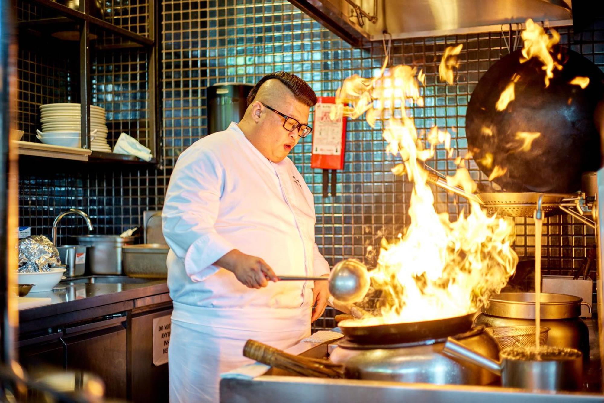 Parkroyal Collection Pickering, Singapura Luaran gambar The photo shows a chef in a kitchen, wearing a white chef's jacket and glasses. He is actively cooking in front of a large wok that is producing a dramatic surge of flames. The kitchen has a modern look, with tiled walls and a variety of kitchen equi