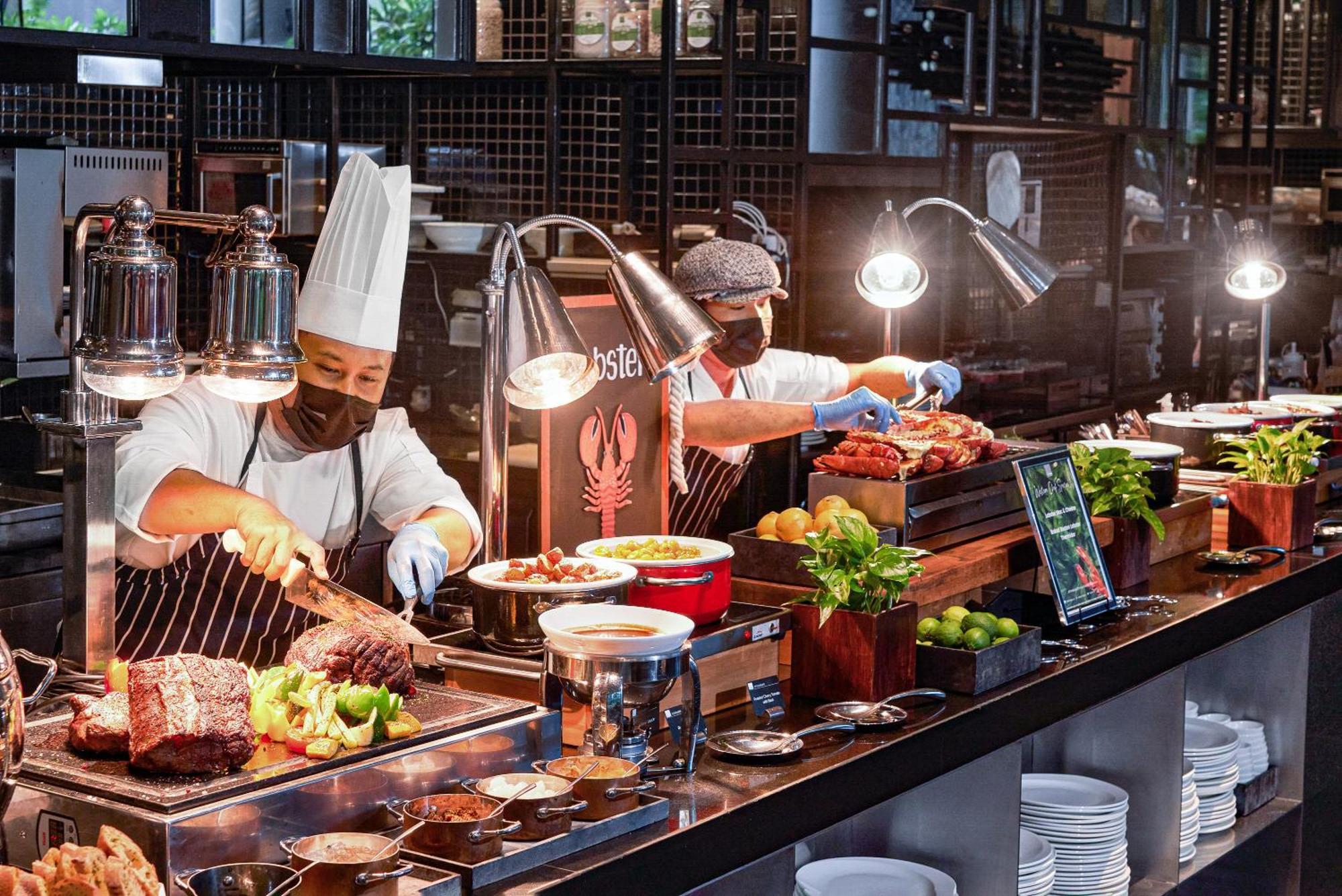 Parkroyal Collection Pickering, Singapura Luaran gambar The photo shows a buffet setup in a restaurant or dining area. In the foreground, two chefs are preparing food. One chef is wearing a white chef's hat and a black mask while slicing meat, and the other is wearing a grey cap and mask, working with var