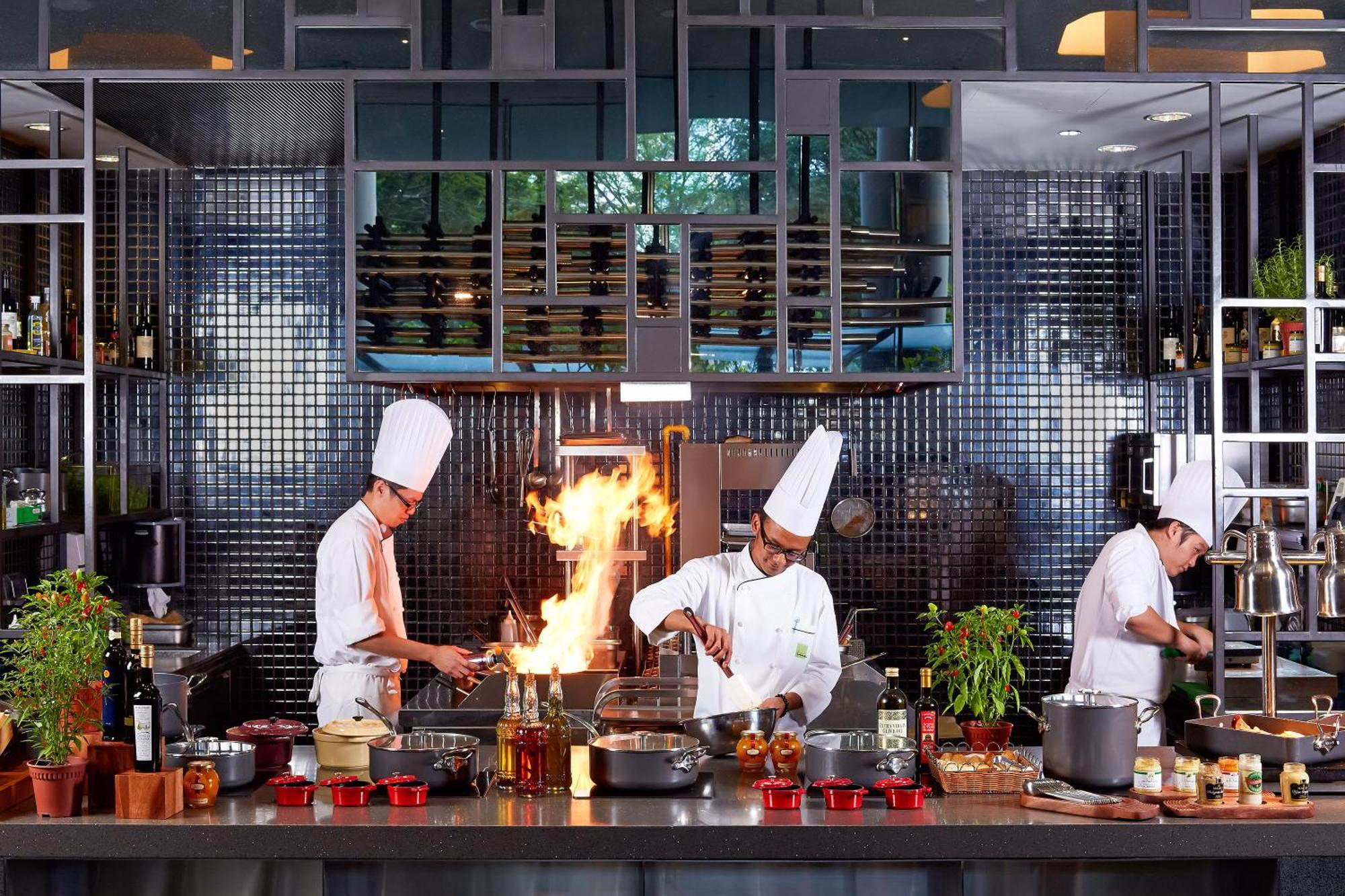 Parkroyal Collection Pickering, Singapura Luaran gambar The photo depicts a modern kitchen where three chefs are actively engaged in cooking. Two of the chefs appear to be stirring pots on the stove, and one chef is working with a flame, likely performing a technique that involves flambéing. The kitchen i