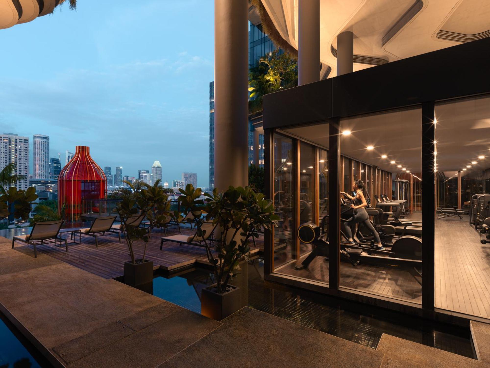 Parkroyal Collection Pickering, Singapura Luaran gambar The photo depicts a modern gym facility that is enclosed with large glass walls, allowing for a view of the outdoors. Inside the gym, there are various exercise machines and equipment. In the background, you can see a city skyline with tall buildings
