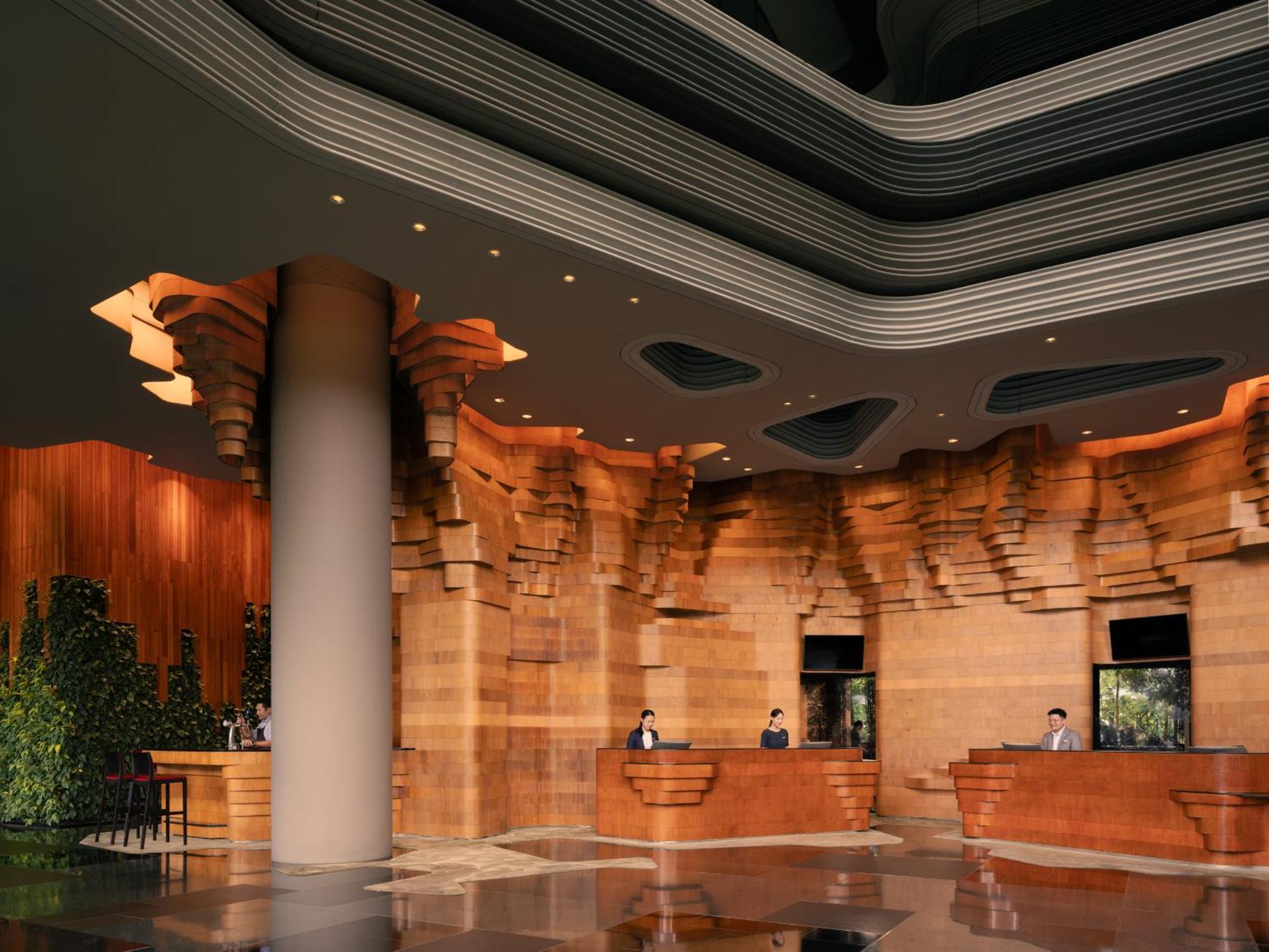 Parkroyal Collection Pickering, Singapura Luaran gambar The image shows a modern hotel or building lobby with a sophisticated design. The interior features richly textured wooden walls that resemble natural formations, creating a warm and inviting atmosphere. The reception area has several service counter