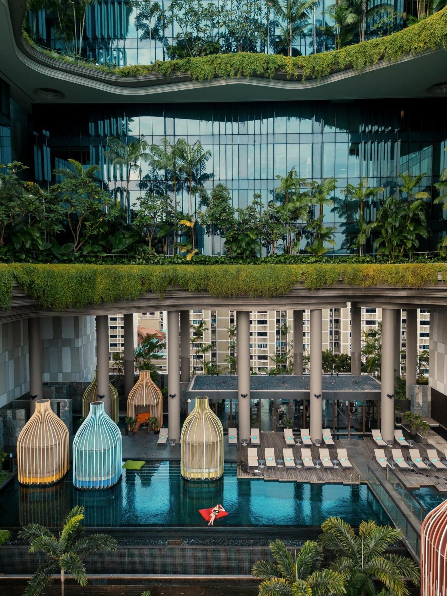 Parkroyal Collection Pickering, Singapura Luaran gambar The photo depicts a modern architectural design featuring a multi-level space. In the foreground, there are colorful structures resembling birdcages, which could be seating areas or decorative elements. Beyond them, there is a pool area lined with su