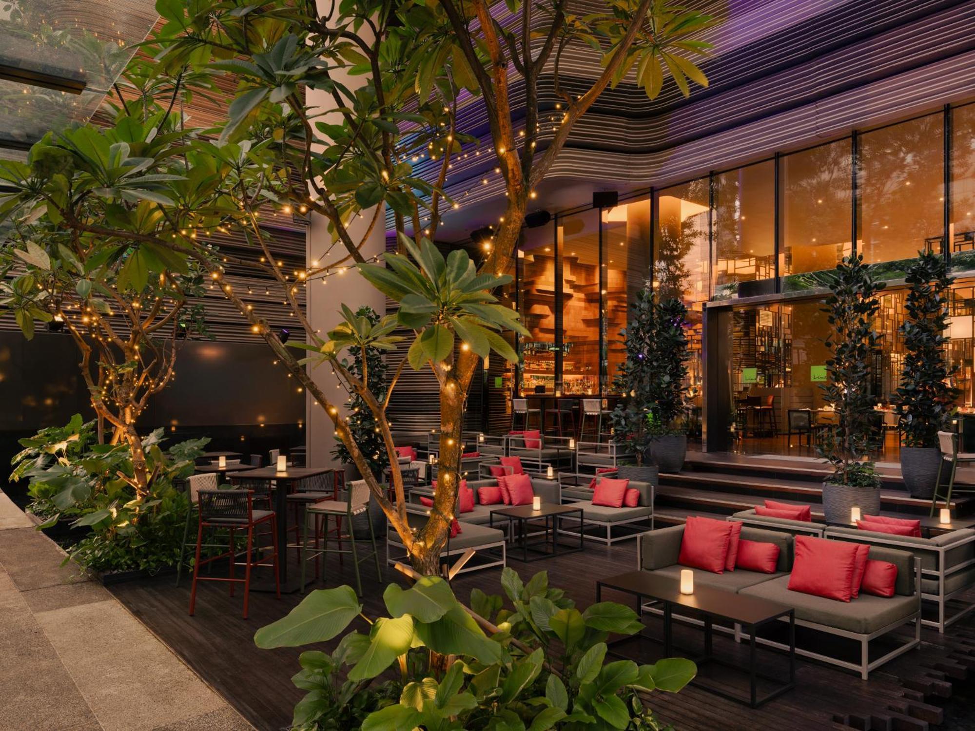 Parkroyal Collection Pickering, Singapura Luaran gambar The photo shows a stylish outdoor seating area of a restaurant or lounge. There are several tables and chairs arranged comfortably, featuring cushions in a vibrant pink color. Surrounding the seating area are lush green plants and trees, which add a 