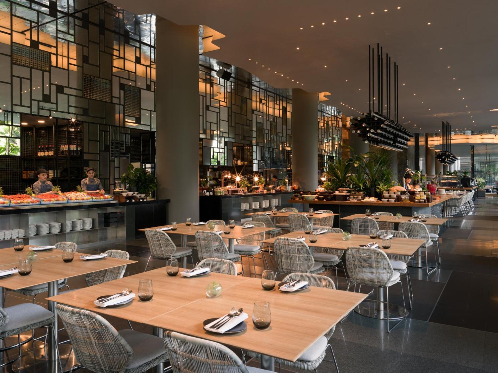 Parkroyal Collection Pickering, Singapura Luaran gambar The photo shows a modern restaurant interior with a spacious layout. There are several tables with wooden tops and transparent chairs arranged neatly. The design features large windows and stylish lighting, creating a warm ambiance. In the background