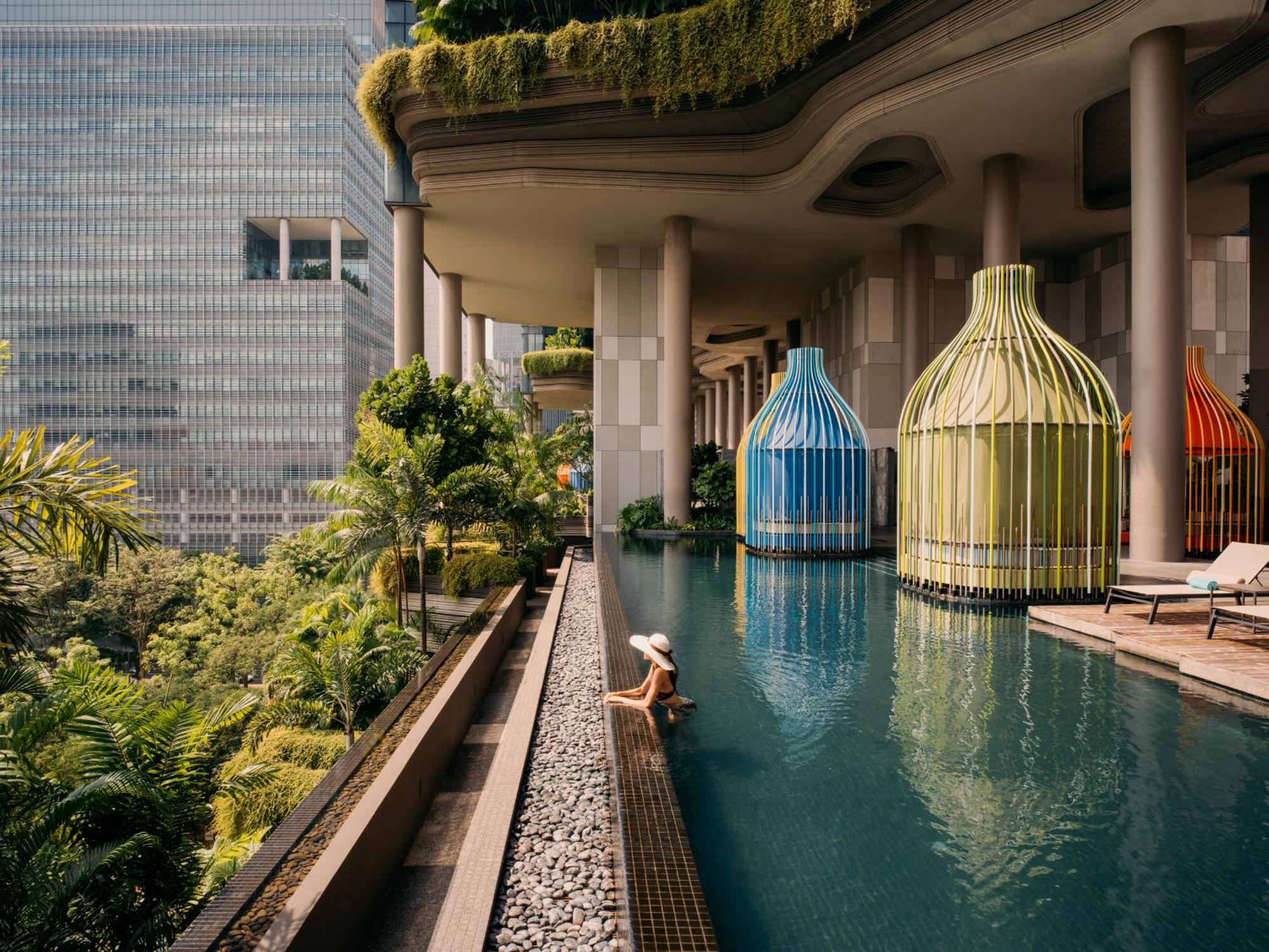 Parkroyal Collection Pickering, Singapura Luaran gambar The photo depicts a beautiful, modern swimming pool area surrounded by lush greenery. There are distinctive, colorful structures resembling large birdcages in blue and green, which add an artistic touch to the scene. A person is seen lounging peacefu